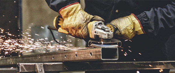 A worker using a handheld angle grinder to cut or smooth a metal beam, with bright sparks flying